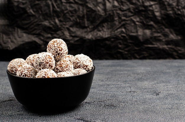 Bolas caseras de dátiles, maní, avena, espolvoreado con coco en un recipiente negro sobre un fondo oscuro con espacio de copia con lugar para el texto.