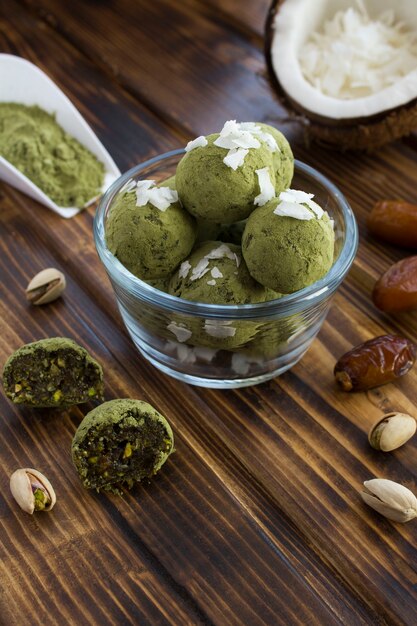 Bolas de energia com pó de matcha, pistache, tâmaras e lascas de coco na tigela de vidro sobre a mesa de madeira marrom.