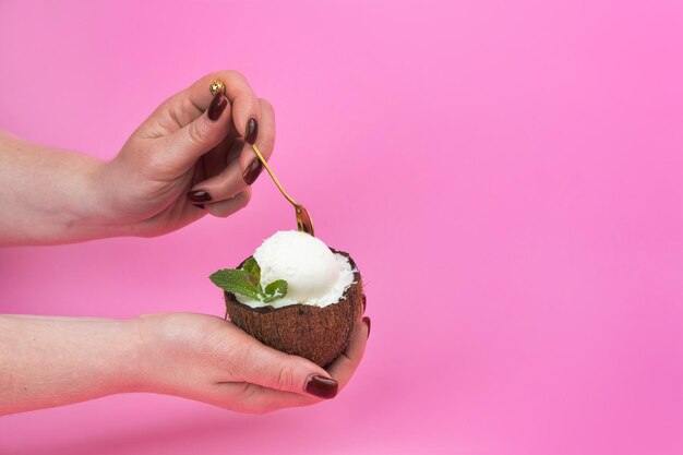 Bolas de helado de vainilla en una mitad de coco fresco, decoradas con hojas de menta en la mano con una cuchara sobre un fondo rosa.