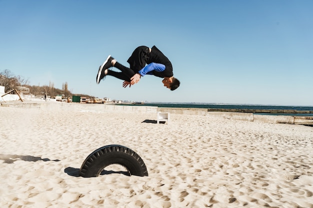 Bold (realce) 20 anos em um agasalho de treino preto fazendo parkour e pulando durante as acrobacias matinais à beira-mar