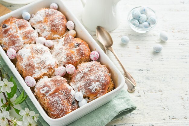 Bolinhos cruzados quentes de Páscoa Tratamentos tradicionais de Páscua bolinhos de cruz com passas, manteiga de chocolate, ovos de doces com flores de cereja ou maçã em fundos rústicos de madeira leve Refeição de férias de Pásqua