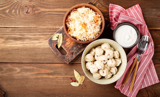 Bolinhos de pelmeni russos tradicionais com creme de leite e chucrute com creme de leite sobre fundo de madeira. Vista superior, configuração plana. Lugar para texto.