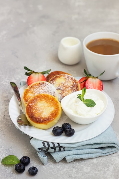 Bolinhos de queijo cottage (syrniki) servidos com creme de leite, frutas frescas (morango e mirtilo) e hortelã. Pequeno-almoço saudável ou almoço de dieta.