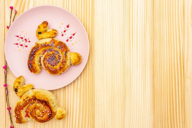 Bollo fresco en forma de un conejito de Pascua. El concepto de panadería de vacaciones para niños. En placa de cerámica rosa con ramas de sauce artificial en superficie de madera, espacio de copia, vista superior.