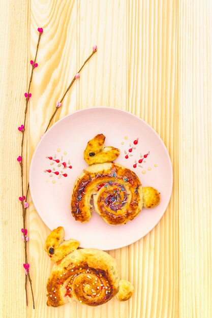 Bollo fresco en forma de un conejito de Pascua. El concepto de panadería de vacaciones para niños. En placa de cerámica rosa con ramas de sauce artificial en la superficie de madera, vista superior, de cerca.