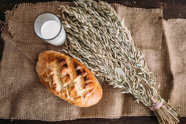 Bollo de pan fresco con corteza crujiente, manojo de trigo y vaso de leche sobre arpillera, vista superior