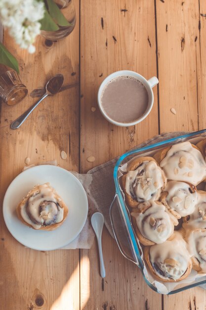 Bollos de canela recién horneados con crema en un plato de vidrio y una taza de cacao en la mesa