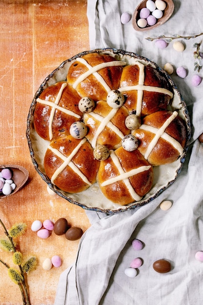 Bollos cruzados calientes tradicionales caseros de Pascua en un plato de cerámica con ramas de sauce en flor y huevos de chocolate caramelos coloridos sobre fondo de piedra naranja. Endecha plana, espacio