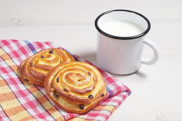 Bollos con pasas y taza de leche esmaltada sobre la mesa de madera clara con mantel