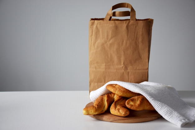 Bollos recién horneados bajo una toalla de cocina blanca sobre una tabla de cortar de madera y un paquete de papel para el servicio de entrega