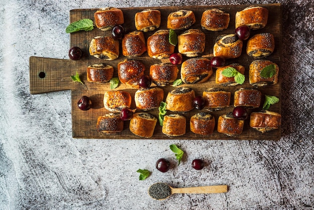 Bollos de semillas de amapola con hojas de menta de cerezas y una cuchara con semillas de amapola en una tabla de madera marrón sobre un fondo gris 1