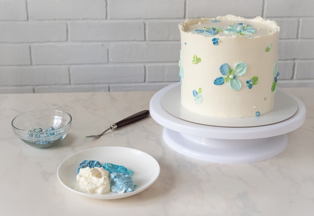 Foto bolo branco alto em um carrinho com flores azuis decoração creme e conjunto para decorar e espátula na mesa.