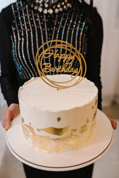 Bolo branco decorado com decoração dourada Bolo de aniversário elegante com topper texto feliz aniversário Aniversariante segurando um bolo nas mãos