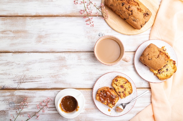 Bolo caseiro com passas, amêndoas, caramelo macio e uma xícara de café em uma superfície de madeira branca e tecido de linho laranja