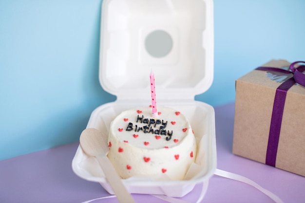 Bolo de aniversário Bento em ecopackaging com uma colher de pau com uma vela ao lado de um presente em um fundo roxo azul