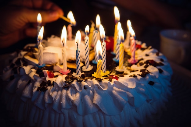 Bolo de aniversário com velas acesas à mão no escuro. Uma mão com um fósforo acende as velas do bolo de aniversário com creme branco. 16 dezesseis velas no bolo.