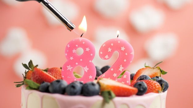Bolo de aniversário número 82 vela rosa no lindo bolo com bagas e isqueiro com fogo contra o fundo de nuvens brancas e céu rosa Vista aproximada