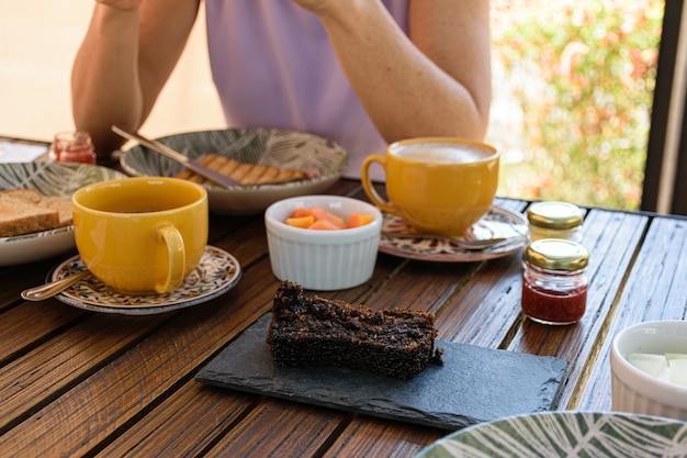 Bolo de biscoito em uma placa de ardósia, ao lado de geleias variadas, mamão, xícara de café com leite e uma mulher.