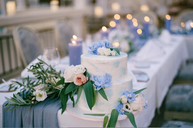 Bolo de casamento de beliche branco decorado com flores e folhas fica em uma mesa com velas