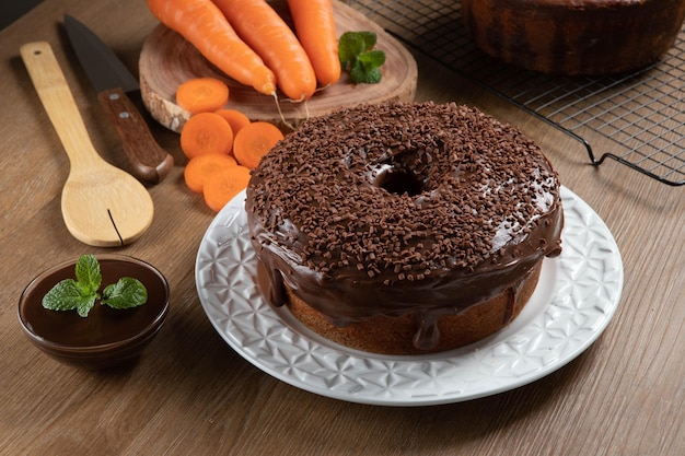 Foto bolo de cenoura brasileiro com cobertura de chocolate na mesa de madeira com cenoura ao fundo