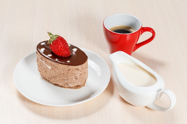 Bolo de chocolate com morango no prato branco, uma xícara de café e uma molheira com creme em uma mesa de madeira