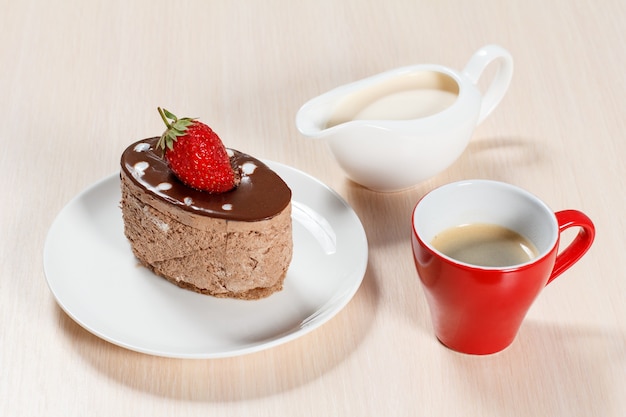 Bolo de chocolate com morango no prato branco, uma xícara de café e uma molheira com leite em uma mesa de madeira