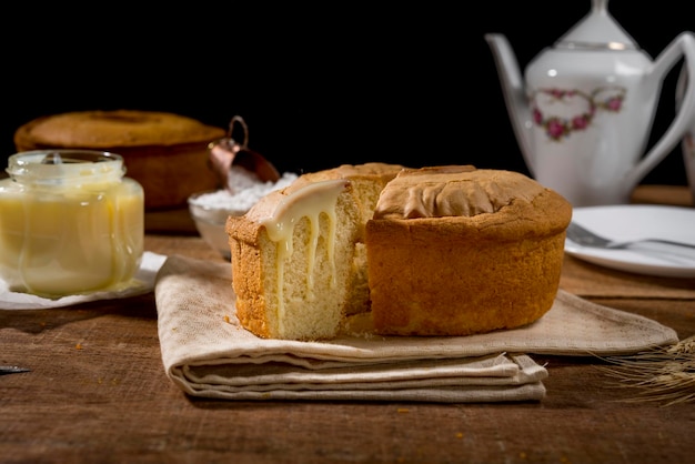 Foto bolo de leite condensado com fatia em madeira rústica e tecido com panela de fermento em pó e bolo no fundo.