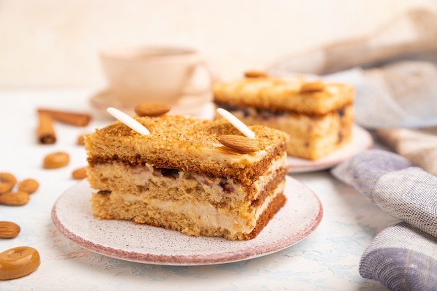 Bolo de mel com creme de leite, caramelo, amêndoas e uma xícara de café em um fundo branco de concreto e têxteis de linho.