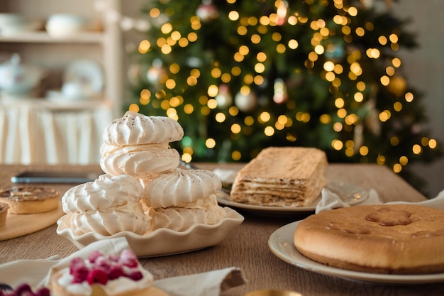 Bolo de merengue branco e arejado, bolo de mel e torta na mesa festiva contra o pano de fundo de uma árvore de natal com uma guirlanda