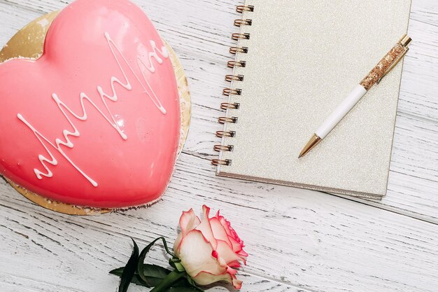 Bolo de mousse rosa em forma de coração na mesa com rosa e bloco de notas