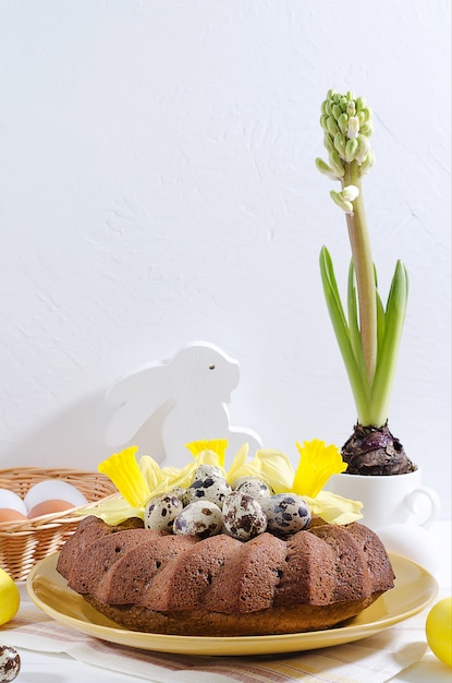 Foto bolo de páscoa com ovos tingidos em um ninho, narcisos e jacinto em um copo em um fundo branco de madeira rústico