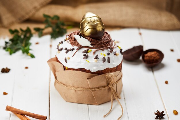 Bolo de Páscoa decorado por ovo de chocolate dourado sobre fundo branco de madeira, tradicional Kulich, Paska, pronto para a celebração