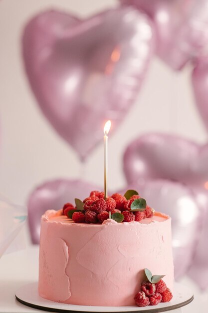 Bolo rosa festivo com framboesas e uma vela no fundo de balões de hélio de corações rosa