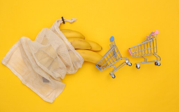 Bolsa de algodón ecológico con plátanos maduros y mini carritos de compra sobre fondo amarillo.