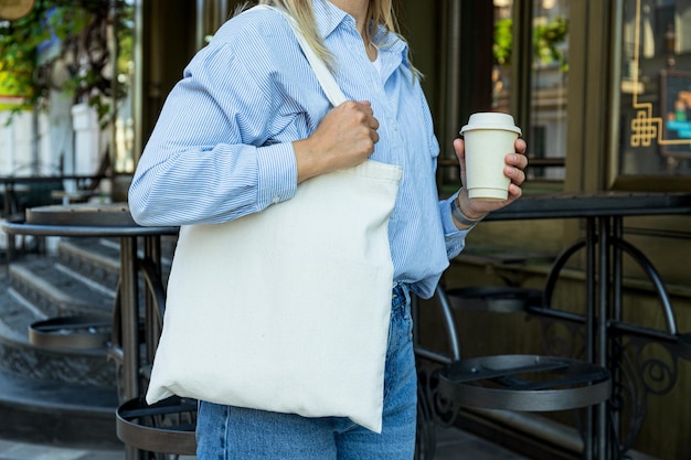 Bolsa blanca de algodón en el hombro de la mujer y taza de papel en la mano al aire libre