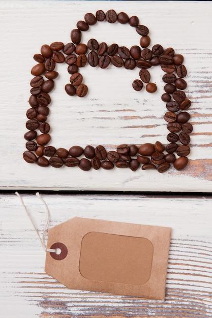 Bolsa com alça feita de grãos de café e etiqueta de papel. Imagem vertical com copyspace. Superfície de madeira branca.