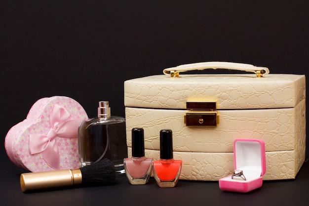 Bolsa de cosméticos, perfume, esmalte de uñas, caja con anillo, pincel y caja de regalo en forma de corazón sobre fondo negro. Concepto de día de celebración.