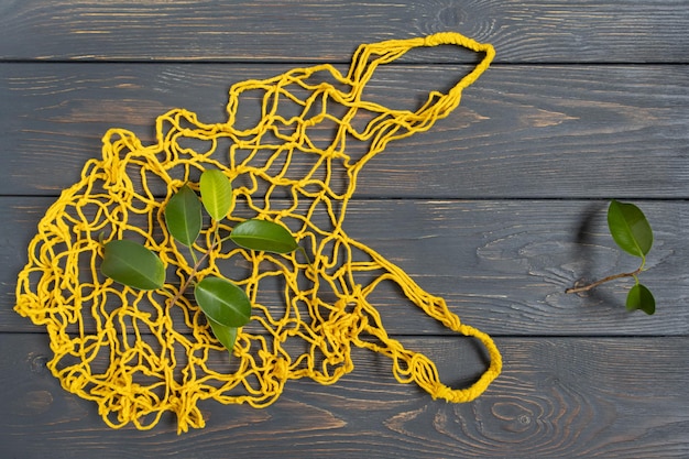 Una bolsa de cuerda tejida amarilla con hojas verdes se encuentra sobre un fondo de madera gris El concepto de reemplazo de bolsas de plástico sin desperdicio
