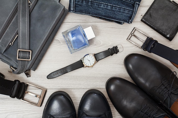 Foto bolsa de ombro, dois pares de sapatos masculinos de couro preto, cintos masculinos, jeans, colônia masculina, relógio de pulso e bolsa em tábuas de madeira cinza. acessórios masculinos