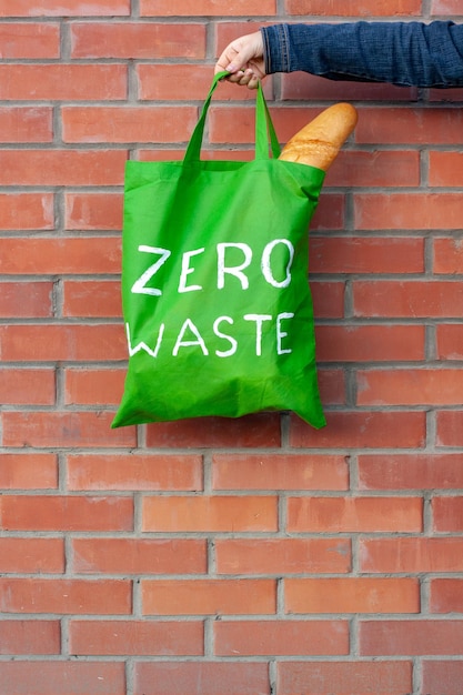 Bolsa ecológica verde con un texto blanco cero desperdicio en la mano sobre un fondo de una pared de ladrillos con espacio de copia. El pan francés es visible en la bolsa. Vertical.