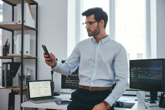 Bolsa. Joven comerciante o hombre de negocios concentrado en ropa formal y anteojos está mirando informes comerciales en su teléfono inteligente con fines de inversión mientras trabaja en su oficina.