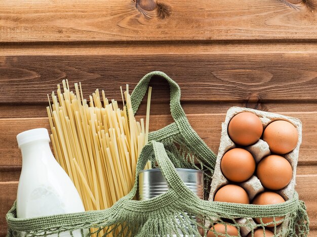 Bolsa de malla de algodón de donación de alimentos con pasta, una lata de comida enlatada, una botella de leche, huevos sobre fondo de madera. Vista superior, endecha plana, espacio de copia.