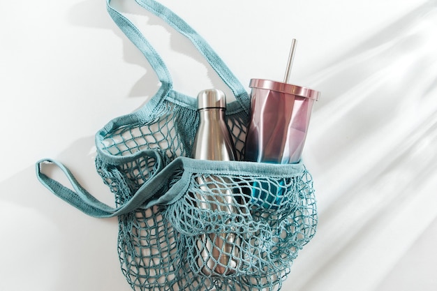 Bolsa de malla con botella de agua reutilizable de metal y taza sobre fondo blanco.