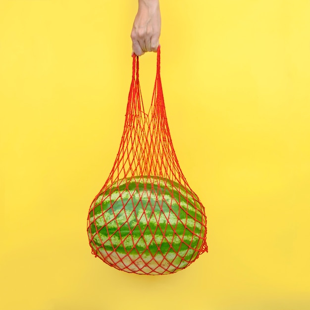 Bolsa de malla con una sandía en mano sobre fondo amarillo
