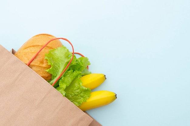 Bolsa de papel con comida: hojas verdes de ensalada, pan y plátano. Disposición sobre fondo azul, vista superior