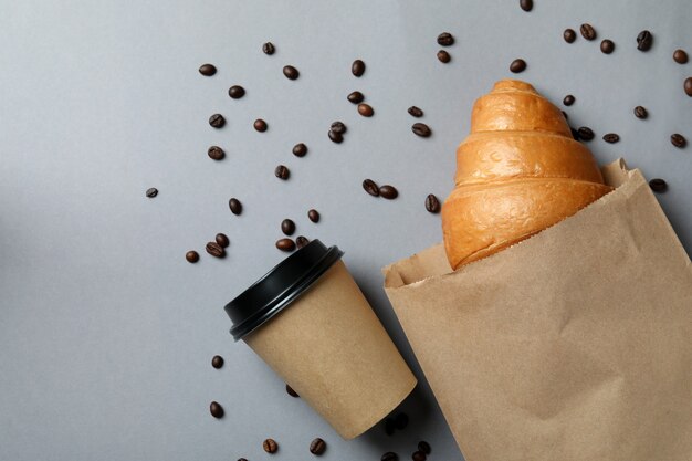 Bolsa de papel con croissant, vaso de papel y semillas de café en gris