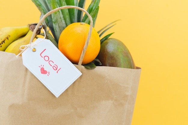 Bolsa de papel con etiqueta con la palabra LOCAL con surtido de frutas sobre fondo amarillo.