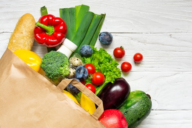 Bolsa de papel llena de diferentes alimentos saludables sobre fondo blanco de madera