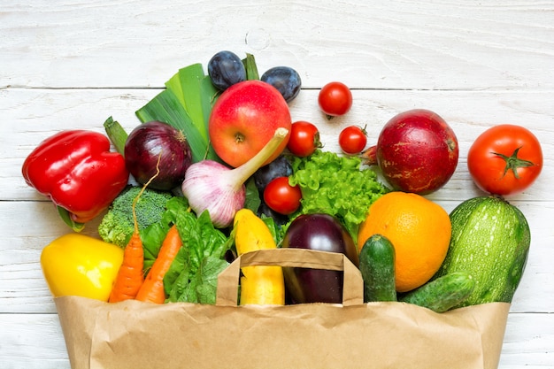 Bolsa de papel llena de diferentes frutas y verduras sobre un fondo blanco de madera