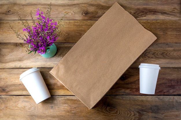 Foto bolsa de papel marrón y dos tazas de papel de café con maqueta de tapas con flores silvestres púrpuras